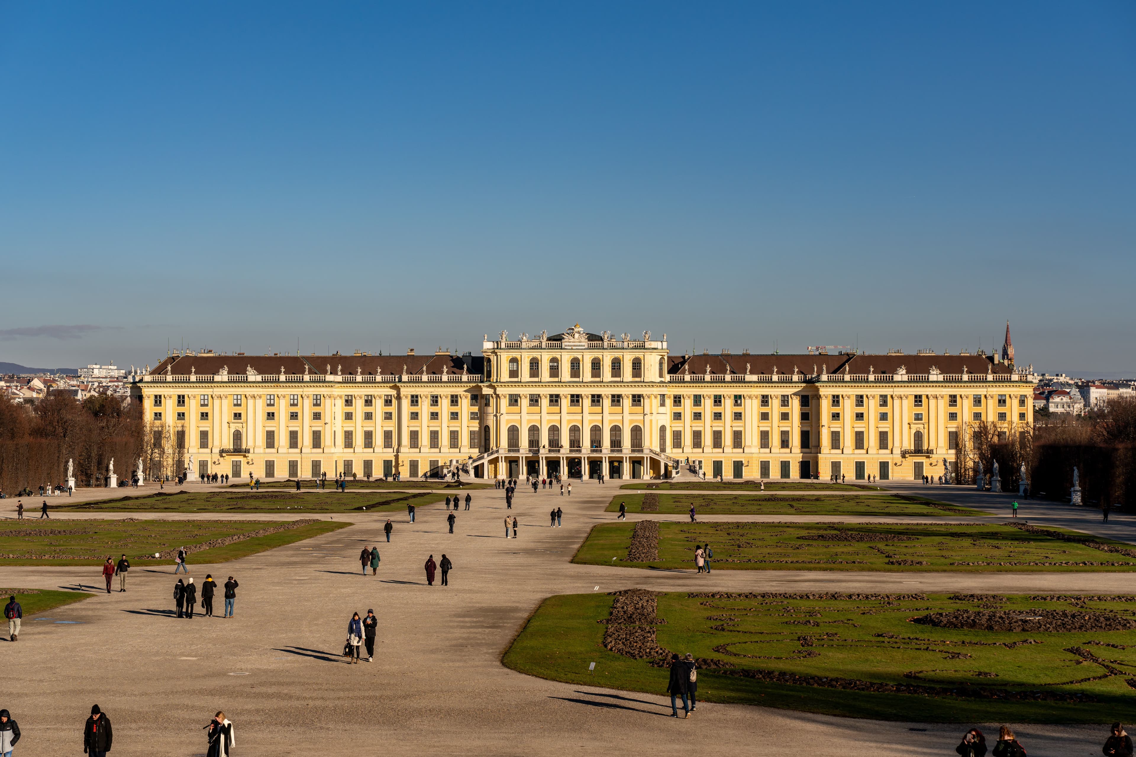 A Schönbrunn kastély sárga homlokzata, előtte a szimmetrikus kastélykerttel és sétáló látogatókkal