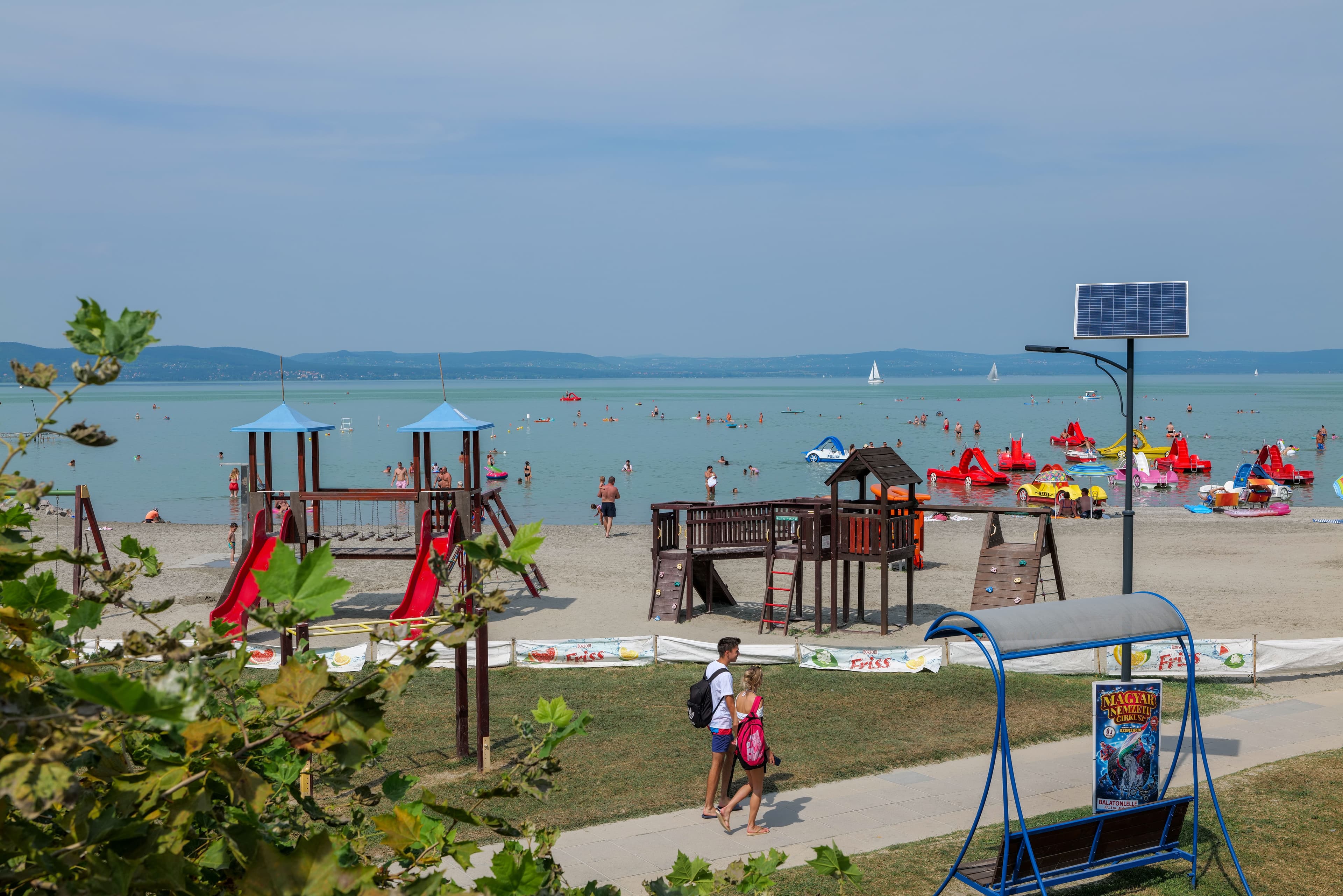 Nyüzsgő homokos Balaton-part játszótérrel, fürdőzőkkel és vízibiciklikkel napsütésben