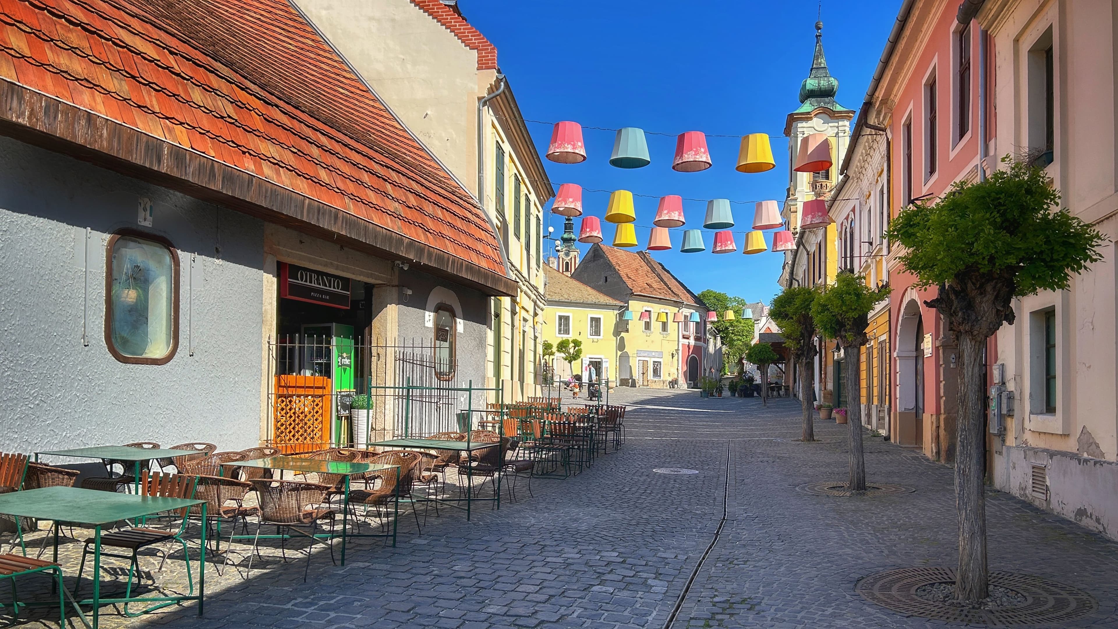 Szentendre hangulatos, macskaköves belvárosi utcája színes lámpaernyő-dekorációval és utcai kávézókkal